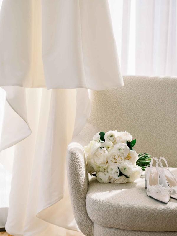 A veil hung up near a white armchair with a bridal bouquet and shoes resting upon it
