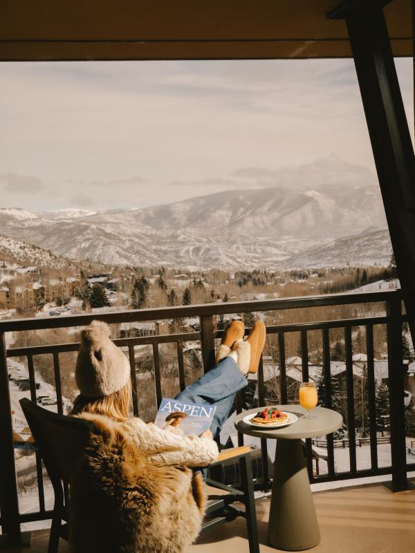 A woman reads a magazine seated in a chair on a balcony overlooking a snowy Colorado mountain range