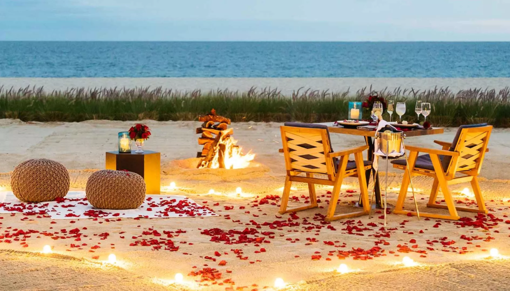 A table with wine and food setup on the beach with a fire and rose pedals laid on the ground at Viceroy Los Cabos