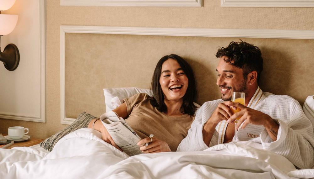 A couple laying in bed with the paper and a mimosa