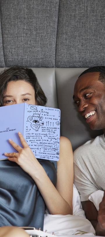 A woman holds a journal up to her face in a bed next to a smiling man holding a coffee cup.