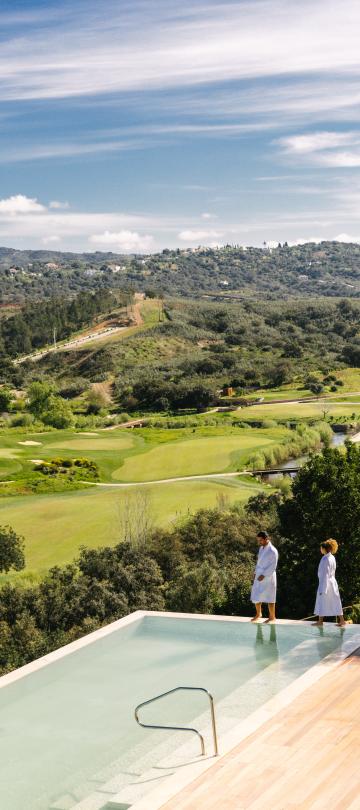 A couple wearing spa bathrobes watches a bird fly over the Viceroy Ombria Algarve golf course, next to a pool