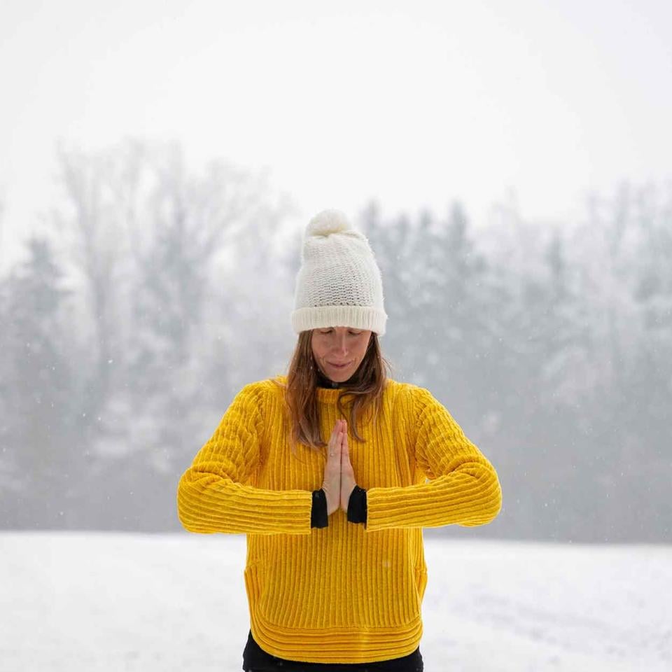 woman in yellow sweater and white hat stands in snowy field with hands in a prayer pose