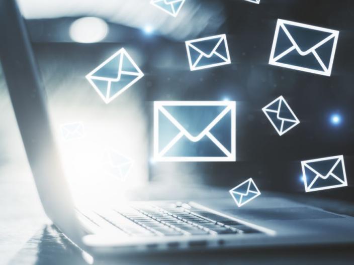 Nine white email icons of various sizes "flying" from the brightness of the open laptop screen. The laptop is facing the right of the image, providing a side profile.