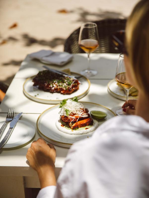 woman wearing white shirt sits at table on beach with plates of food