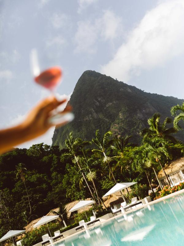 person holding wine glass in front of pool, cabanas, and mountain