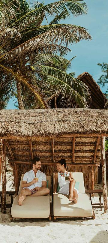 A man and woman sitting on cabanas under palm trees on the beach
